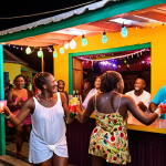 A vibrant, authentic street bar in Basseterre, St. Kitts at night. Locals and tourists laughing and dancing spontaneously to a live reggae and calypso band. Colorful strung lights illuminate the festive atmosphere, with people holding rum punch cocktails, capturing the joyful communal spirit.